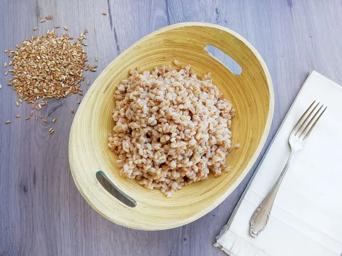 Farro integrale cotto in una ciotola di legno, pronto per gustose ricette. Accanto, chicchi di farro crudo e una forchetta.