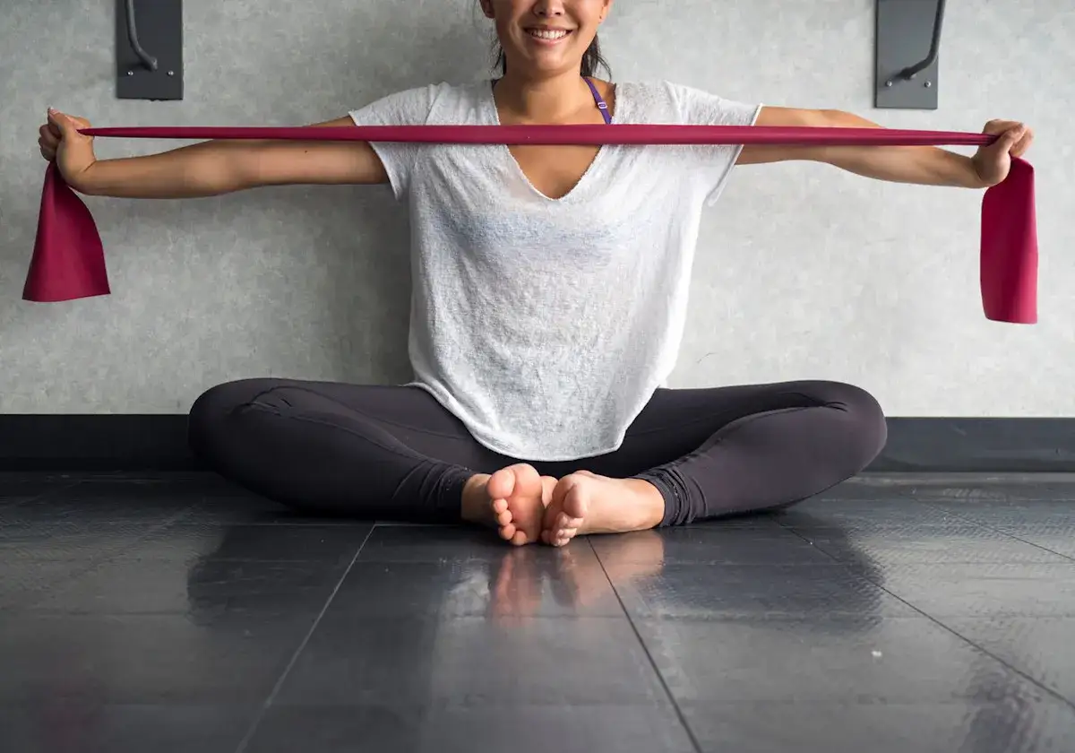 Donna sorridente esegue esercizi con elastici, seduta a gambe incrociate sul pavimento.