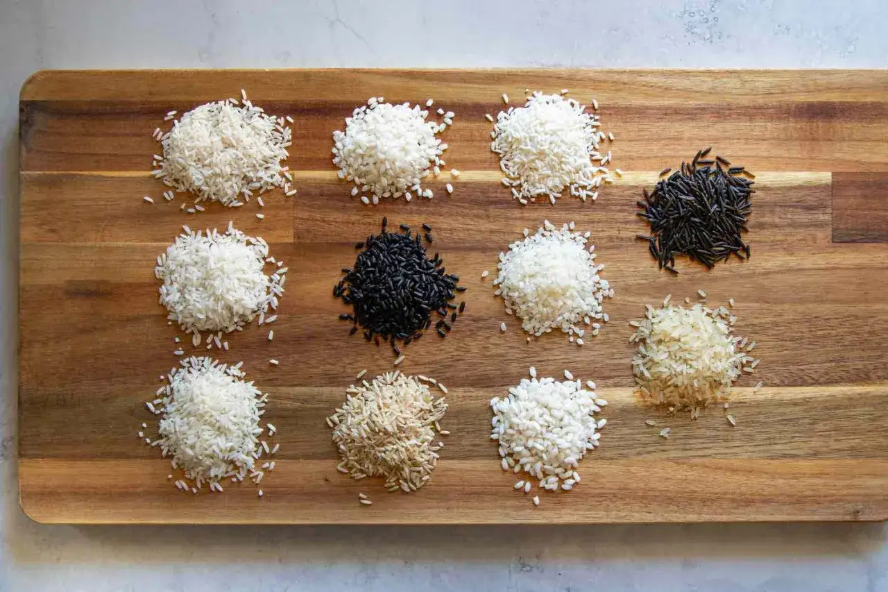 Nove mucchietti di riso di diverse variet&agrave;, dal bianco al nero, disposti su un tagliere di legno. Un invito a scegliere il tuo riso pi&ugrave; leggero per ogni piatto.