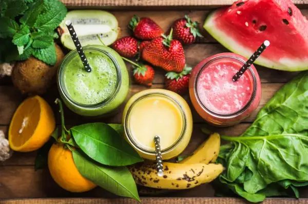 Tre colorati frullati di frutta con acqua, circondati da ingredienti freschi come fragole, kiwi, arance e spinaci.