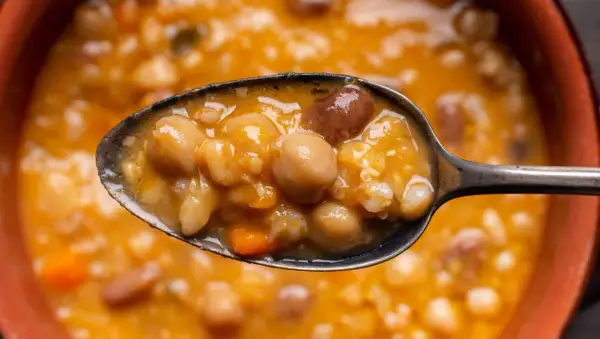 Un cucchiaio pieno di zuppa ricca di legumi e cereali, con ceci, fagioli e pezzetti di carota. Un piatto confortante e nutriente.