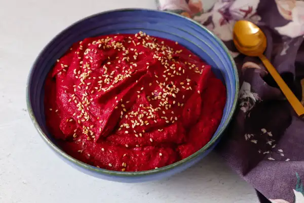 Hummus di barbabietola cremoso e vibrante, cosparso di semi di sesamo, in una ciotola blu. Un cucchiaio dorato e un tovagliolo fiorato completano la scena.