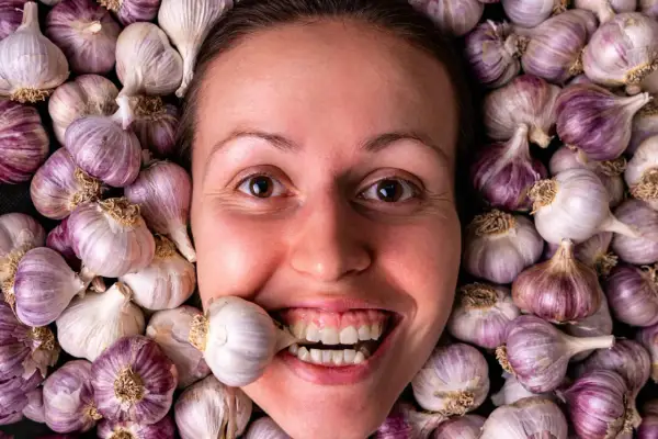 Una donna sorride con uno spicchio d'aglio tra i denti, circondata da teste d'aglio. Si dice che l'aglio fa dimagrire!
