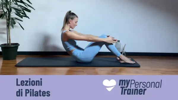 Donna esegue un esercizio di pilates matwork, seduta sul tappetino con le ginocchia piegate e le braccia tese in avanti.