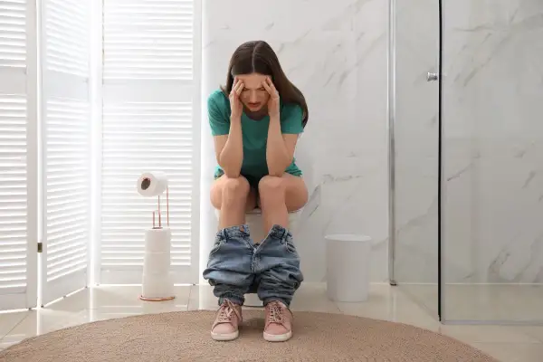 Donna con mani sulla testa, seduta sul water, con jeans abbassati. Sembra soffrire, forse ha mangiato troppo e ora feci caprine.
