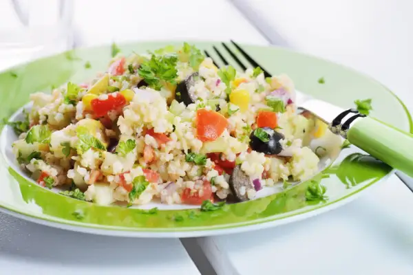Un'insalata di miglio colorata con pomodori, olive, mais e prezzemolo fresco, servita su un piatto verde brillante.